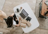 From above of young woman with long dark hair in casual clothes working at table and browsing netbook while sitting in modern workplace and touching hair
